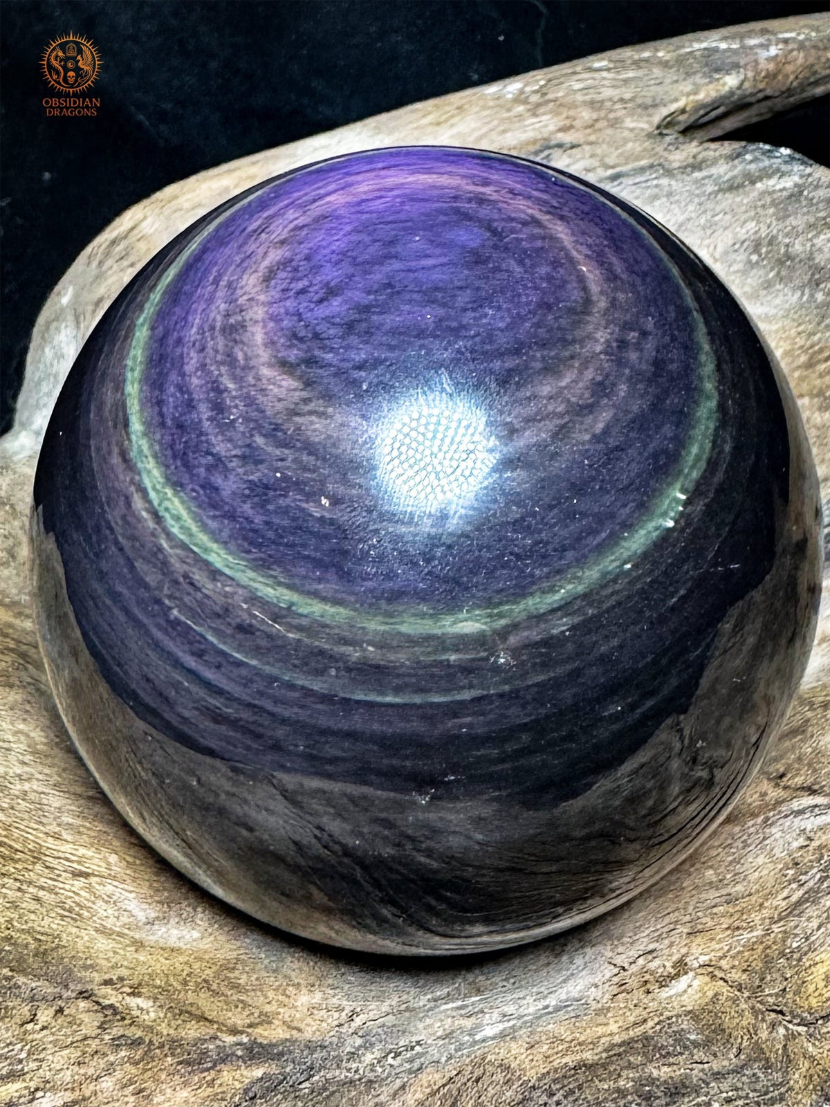 Sphère en obsidienne Manta Huichol de 7 cm inclinée, grande plage violette et lavande bordée de vert au-dessus de bandes sombres.