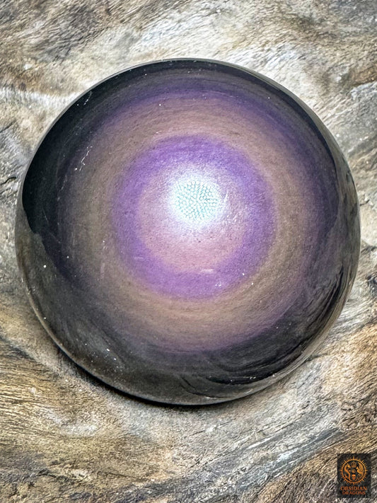 Sphère en obsidienne œil céleste aux reflets violets, cœur mauve rosé et anneau violet soutenu, posée sur bois - Obsidian Dragons