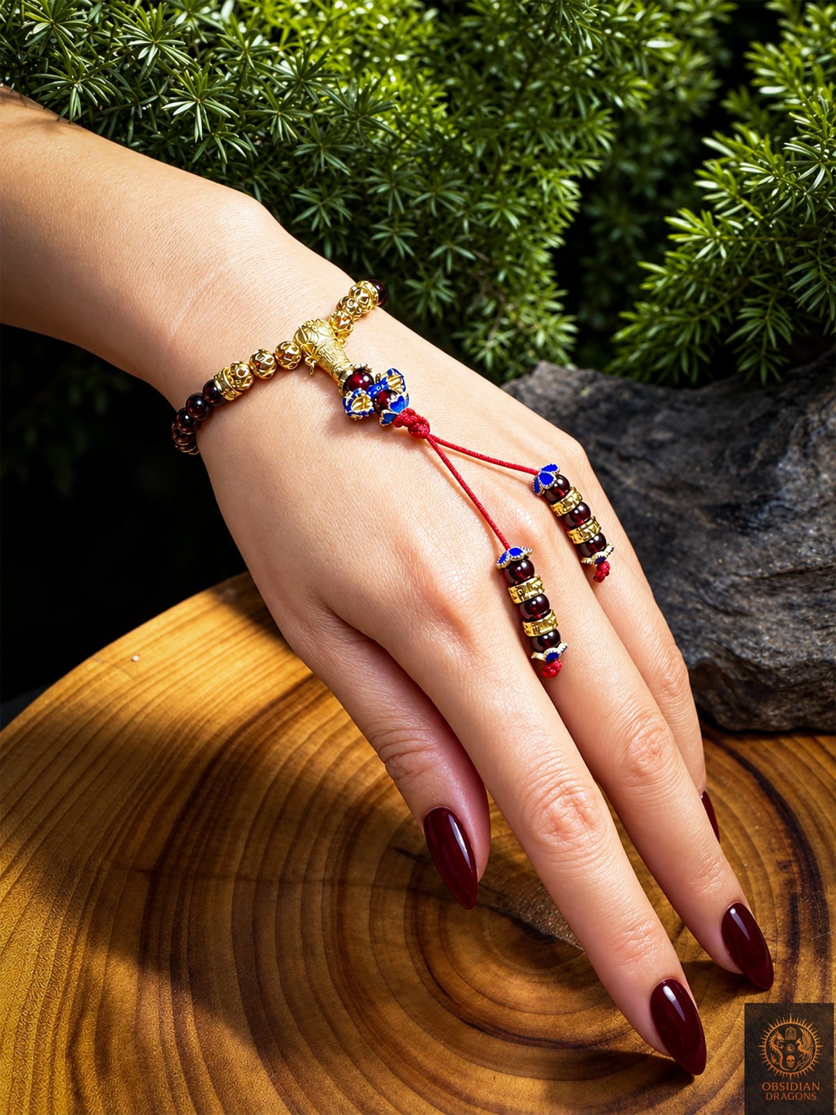 Bracelet mala en grenat porté au poignet, avec perle de Guru dorée, dorje cloisonné bleu et deux brins rouges ornés de grenats sur la main - Obsidian Dragons