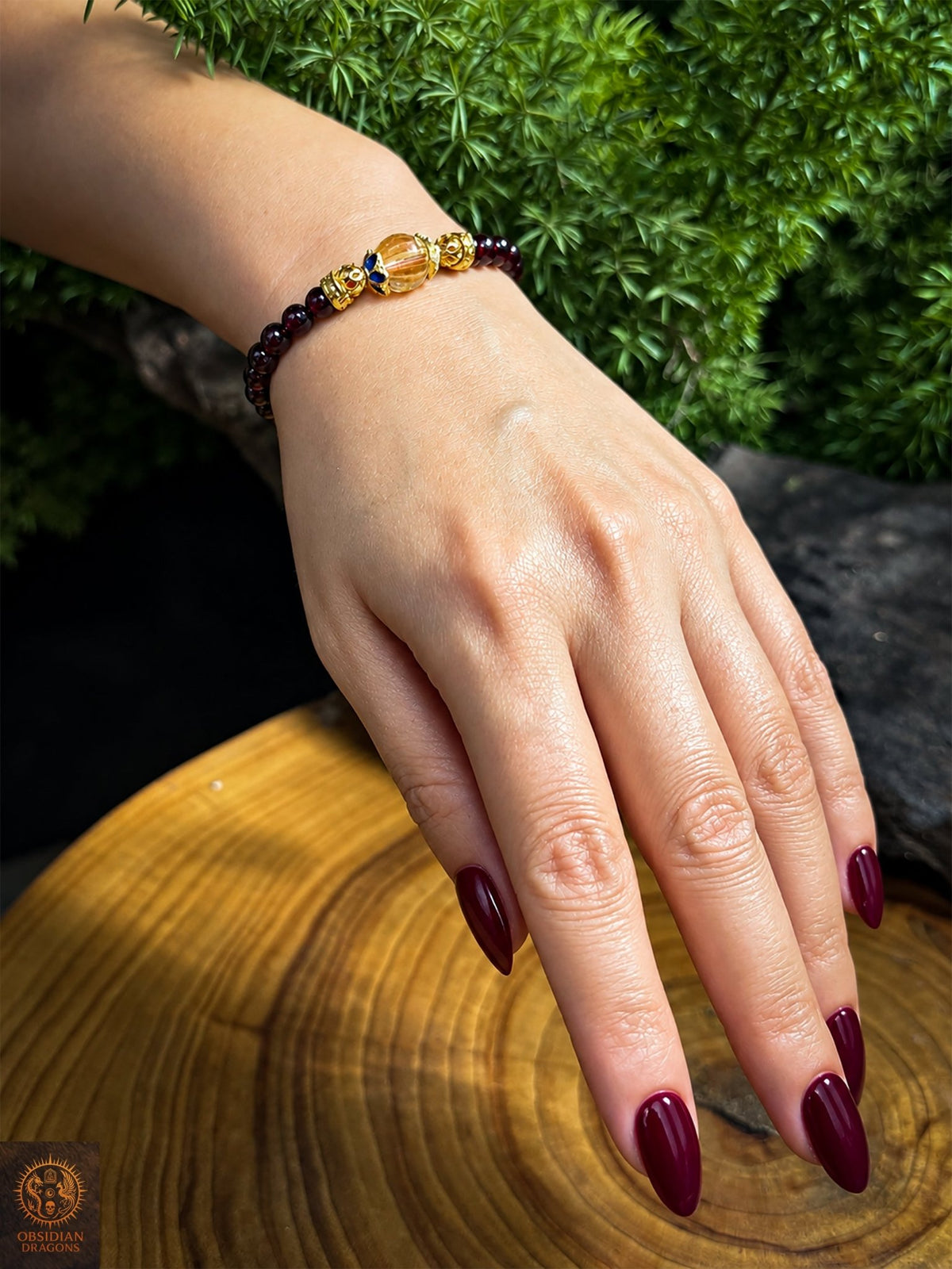 Bracelet mala en grenat et citrine porté au poignet, avec perle centrale jaune facettée et fleurs émaillées bleues en bronze plaqué or - Obsidian Dragons