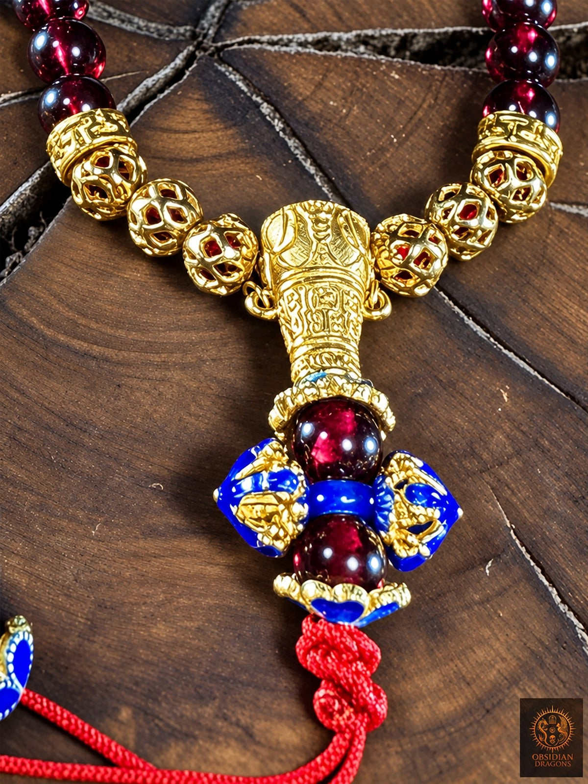 Gros plan sur la perle de Guru en bronze plaqué or et le dorje cloisonné bleu du bracelet mala en grenat, avec son nœud rouge.