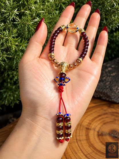 Bracelet mala en grenat et citrine présenté dans la paume, avec dorje cloisonné et deux brins de cordon rouge terminés par des grenats.