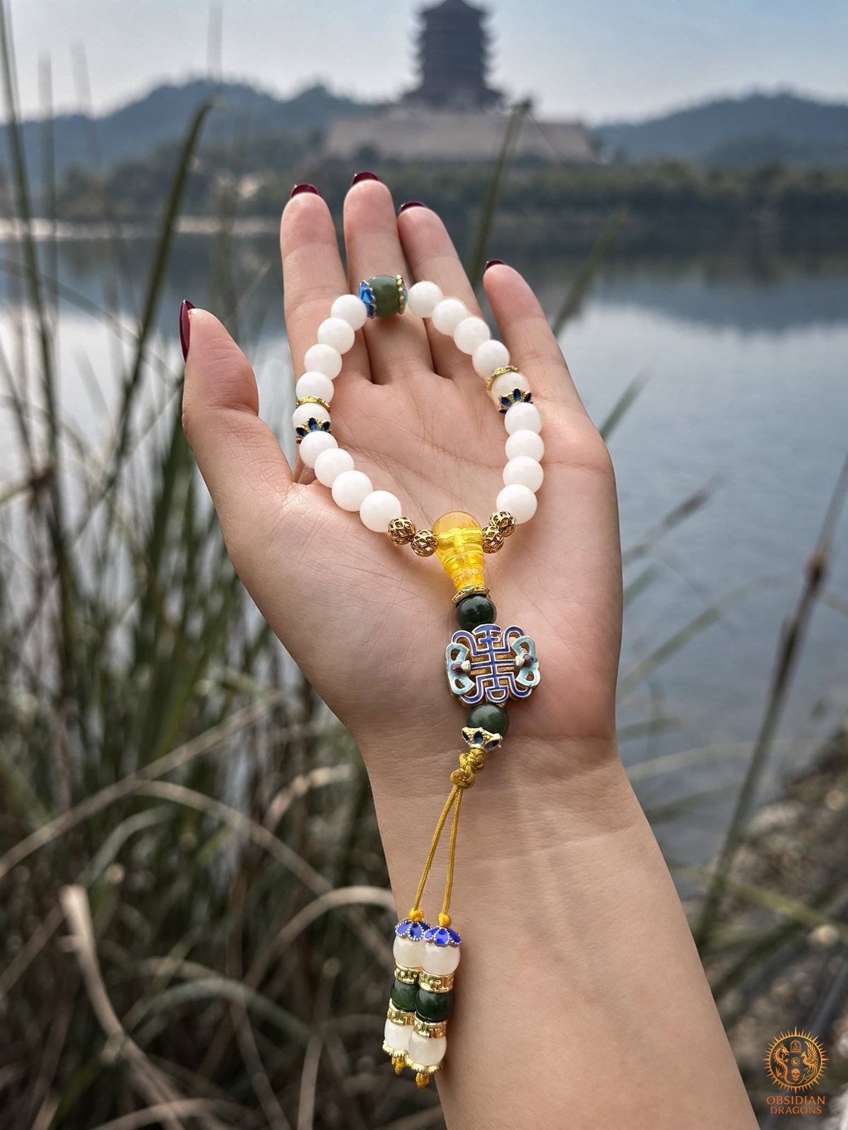 Shibazi de 18 perles en jade blanc présenté dans une paume, avec perle verte, ambre et motif shòu devant un lac.