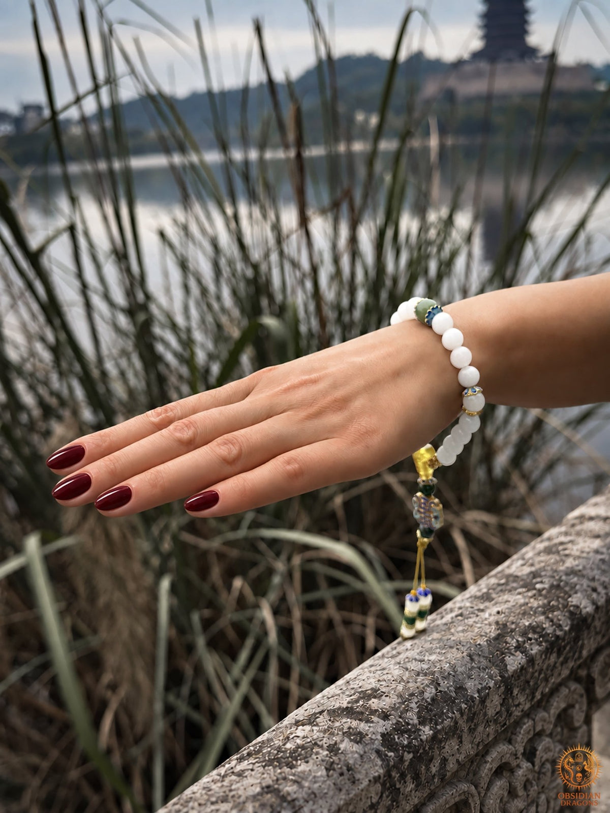 Bracelet shibazi en jade blanc porté au poignet, avec perle verte et ornements suspendus, photographié au bord d’un lac.