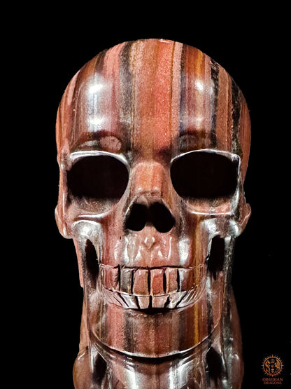 Vue de face d’un crâne en œil de fer sculpté à la main, aux orbites profondes et aux bandes minérales rouges et sombres - Obsidian Dragons