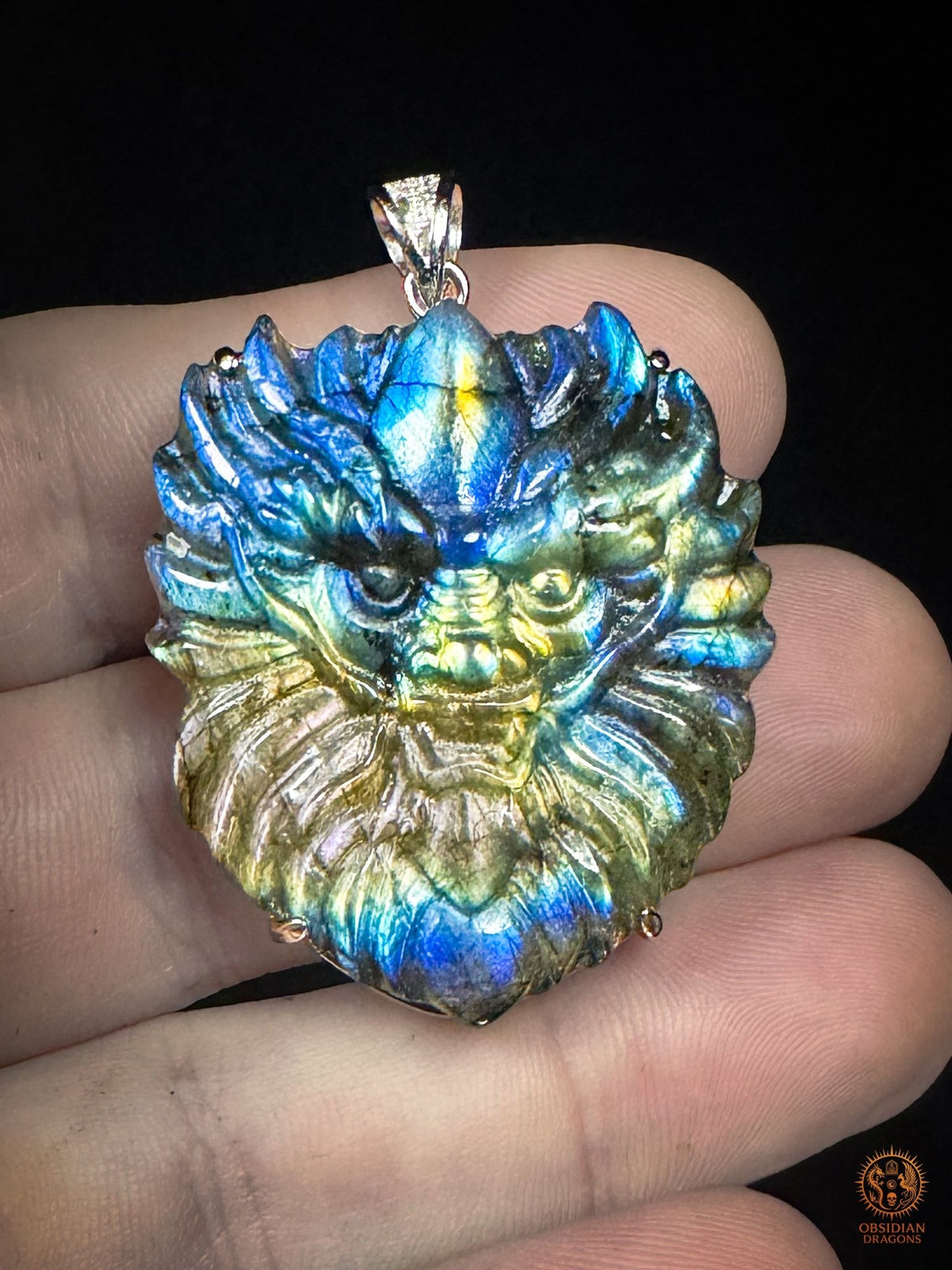 Pendentif dragon en labradorite tenu en main, montrant ses dimensions, sa sculpture détaillée et ses reflets bleus, verts et dorés