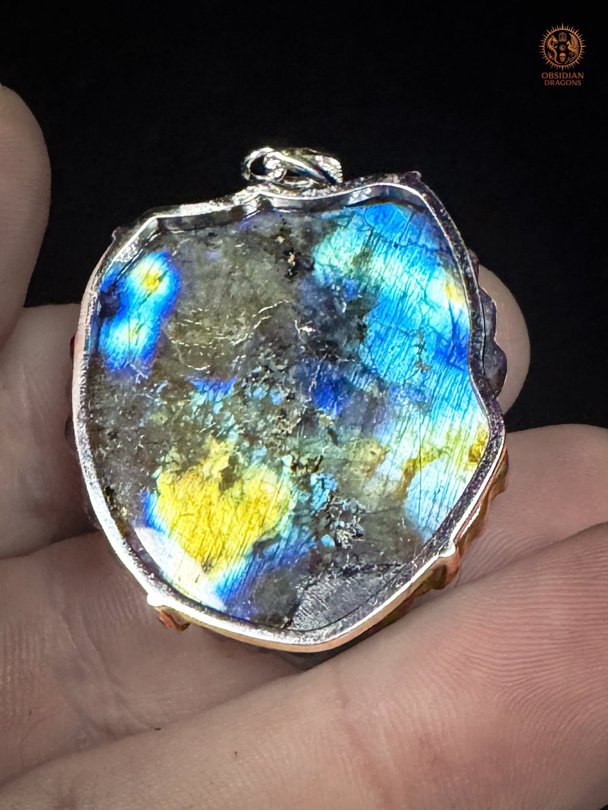 Revers du pendentif dragon en labradorite tenu en main, révélant la pierre naturelle et sa labradorescence bleue, turquoise et dorée