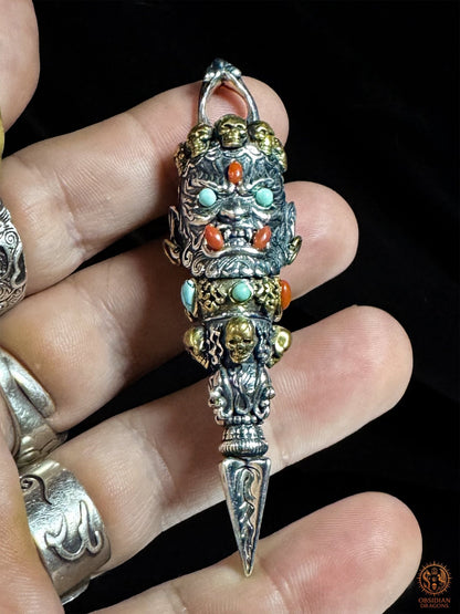 Gros plan d’un pendentif Phurba Mahakala tenu entre les doigts, dague rituelle tibétaine en argent 925 et bronze avec pierres naturelles
