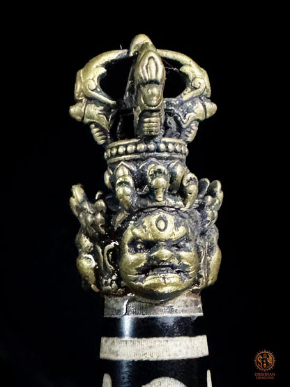 Gros plan sur la poignée du Phurba en bronze ornée de visages courroucés et surmontée d’un vajra
