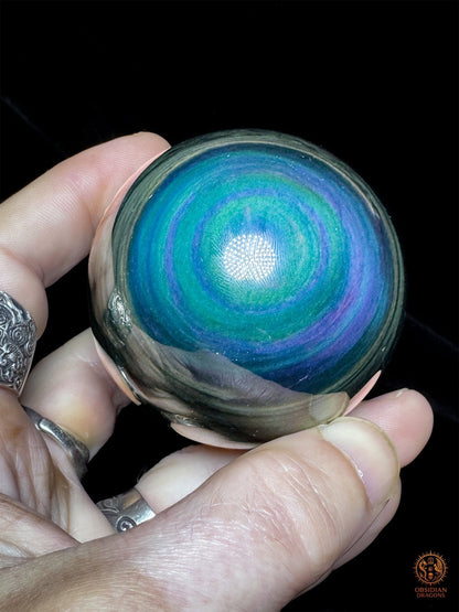 Sphère en obsidienne Manta Huichol tenue en main, montrant ses irisations bleu cyan, turquoise et violettes sur fond noir - Obsidian Dragons