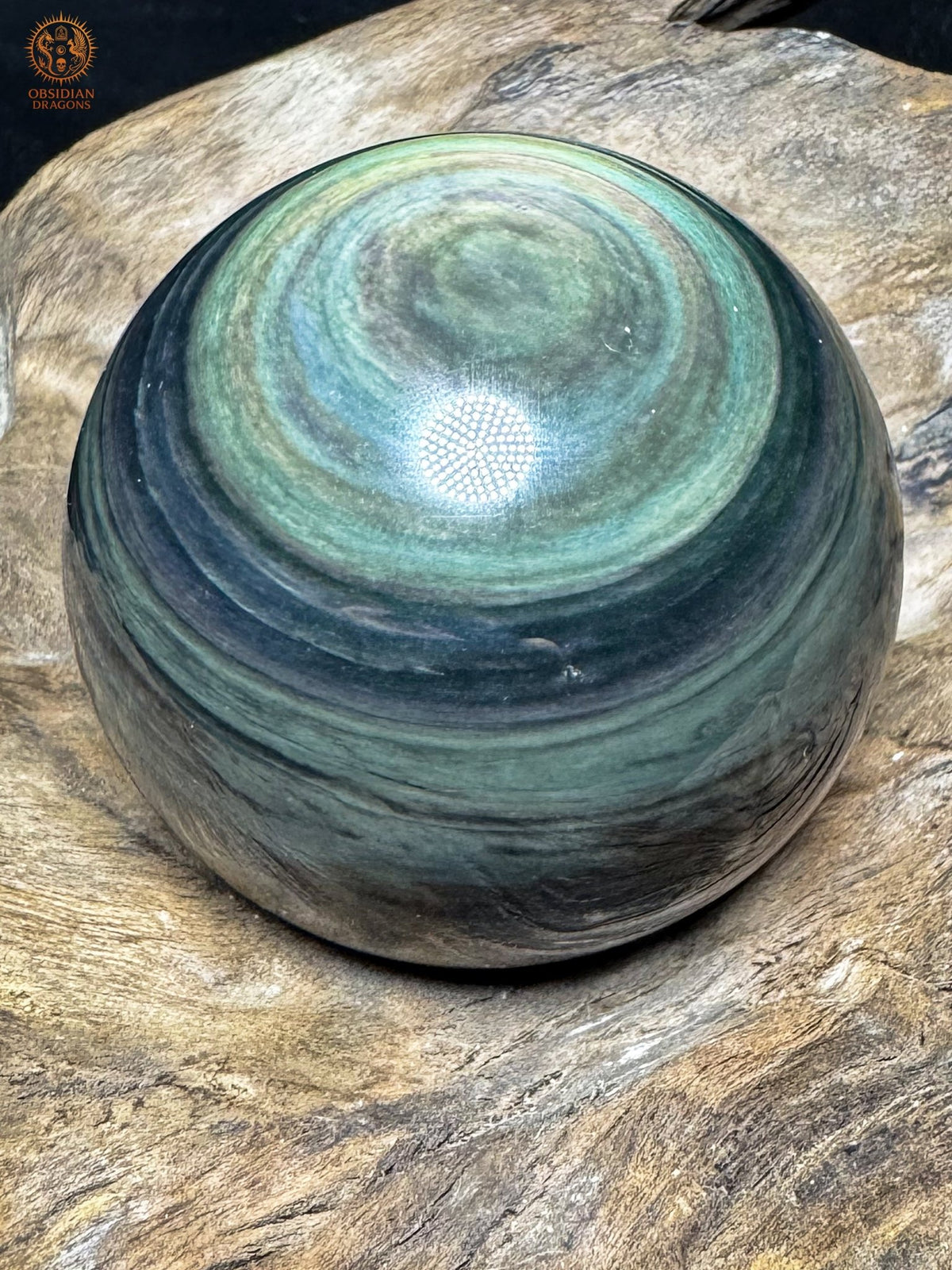 Sphère en obsidienne Manta Huichol inclinée, œil vert pâle en partie haute et rubans gris-vert sur les côtés sombres.