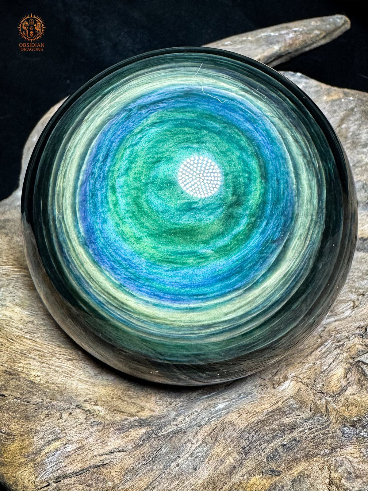 Sphère en obsidienne Manta Huichol de 8,12 cm, avec un centre vert entouré de rubans bleus et jaune-vert, posée sur bois - Obsidian Dragons