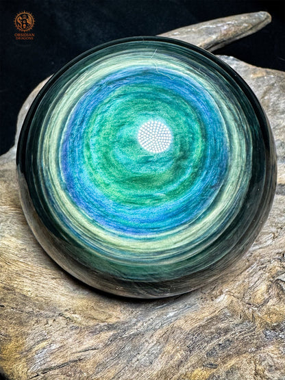 Sphère en obsidienne Manta Huichol de 8,12 cm, avec un centre vert entouré de rubans bleus et jaune-vert, posée sur bois - Obsidian Dragons
