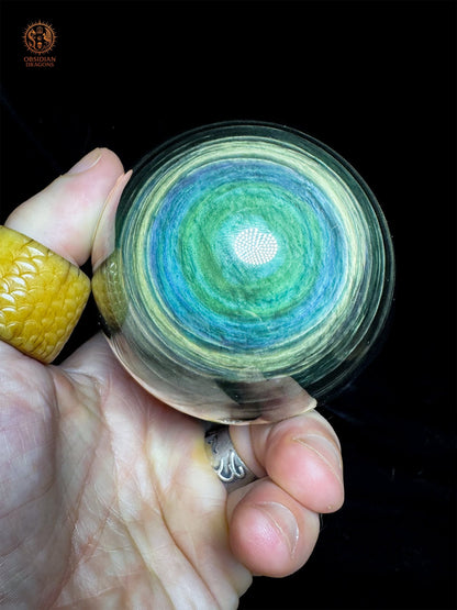 Sphère en obsidienne Manta Huichol de 8,12 cm tenue en main, révélant un œil vert bordé de bleu et de couronnes jaune-vert - Obsidian Dragons