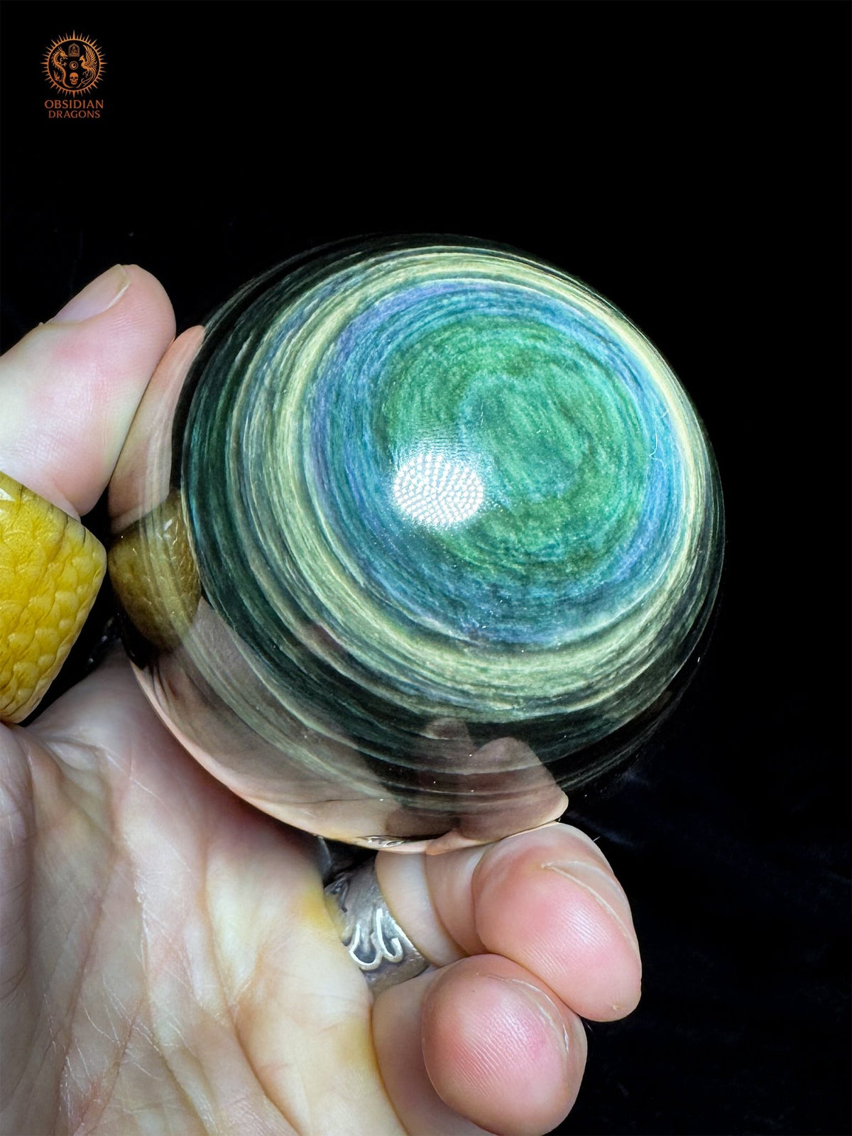 Sphère en obsidienne Manta Huichol tenue en main et inclinée, aux bandes fines bleues et jaune-vert autour d’un large centre vert - Obsidian Dragons
