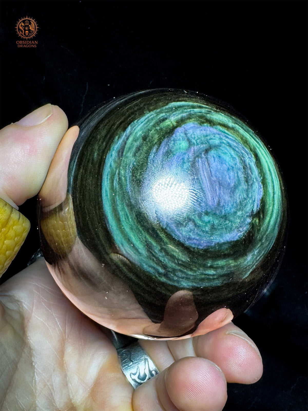 Vue oblique de la sphère en obsidienne Manta Huichol tenue en main, montrant des volutes bleu lavande et des couronnes vertes - Obsidian Dragons