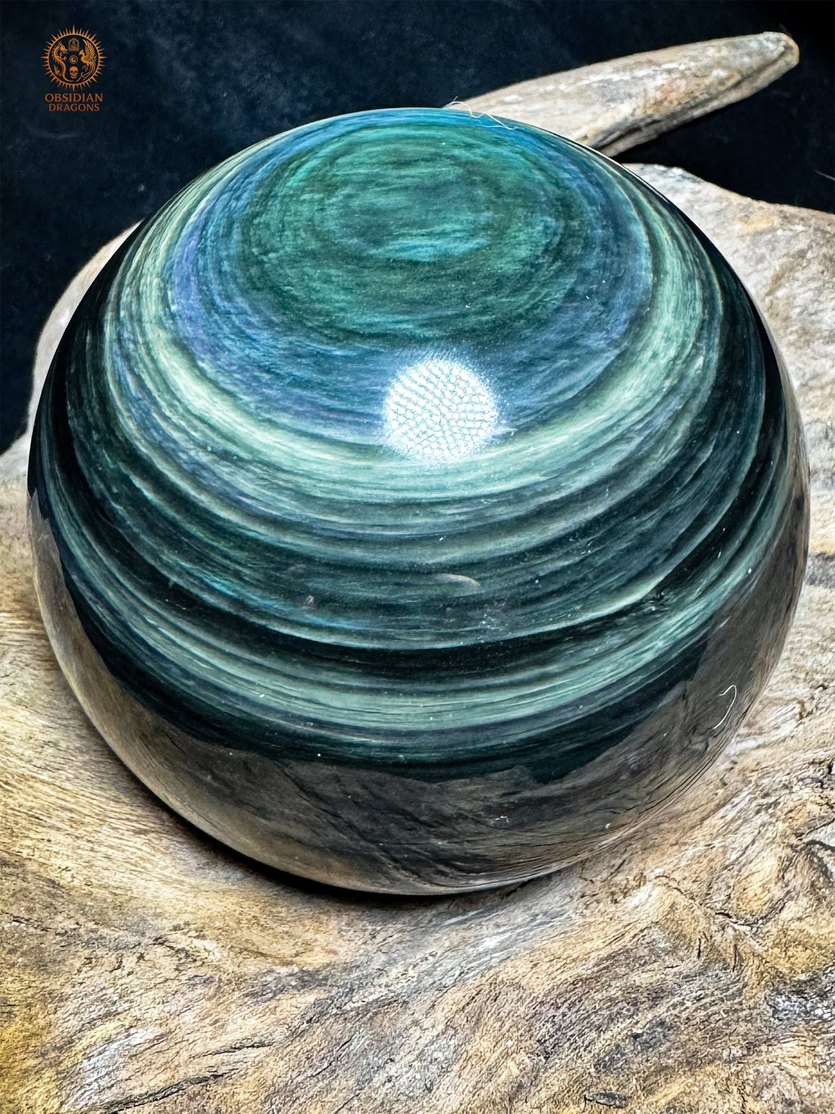 Sphère en obsidienne Manta Huichol posée sur bois, avec un œil vert en partie haute et de nombreux rubans bleus et vert pâle.