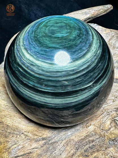 Sphère en obsidienne Manta Huichol posée sur bois, avec un œil vert en partie haute et de nombreux rubans bleus et vert pâle.