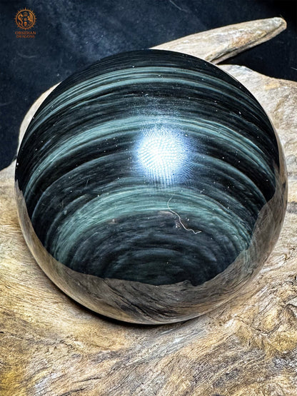 Vue latérale de la sphère en obsidienne œil céleste, aux fines bandes bleu-gris ondulantes sur une surface noire polie.