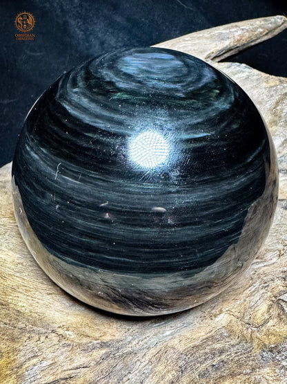 Sphère en obsidienne polie posée sur bois, avec un halo bleu décentré vers le haut et de fines bandes irisées sur fond sombre.