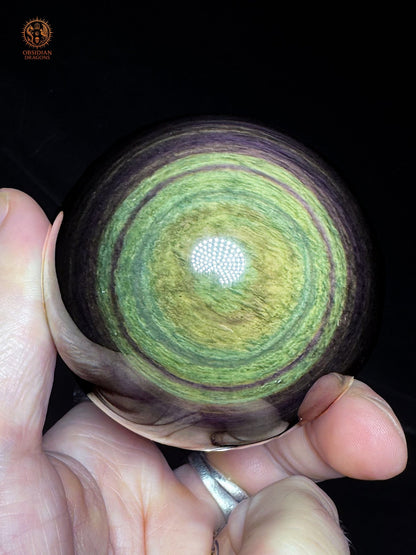Sphère en obsidienne Manta Huichol de 7 cm tenue en main, montrant ses reflets jaune-vert et ses couronnes vertes et violettes - Obsidian Dragons