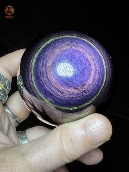 Obsidienne œil céleste Manta Huichol tenue en main, centre bleu-violet entouré de bandes lavande et d’un anneau vert clair - Obsidian Dragons
