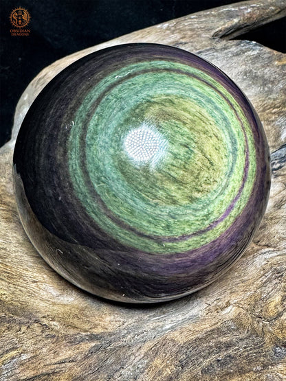 Sphère en obsidienne Manta Huichol inclinée sur bois, centre jaune-vert décentré, bandes vert d’eau et contours violets panachés.