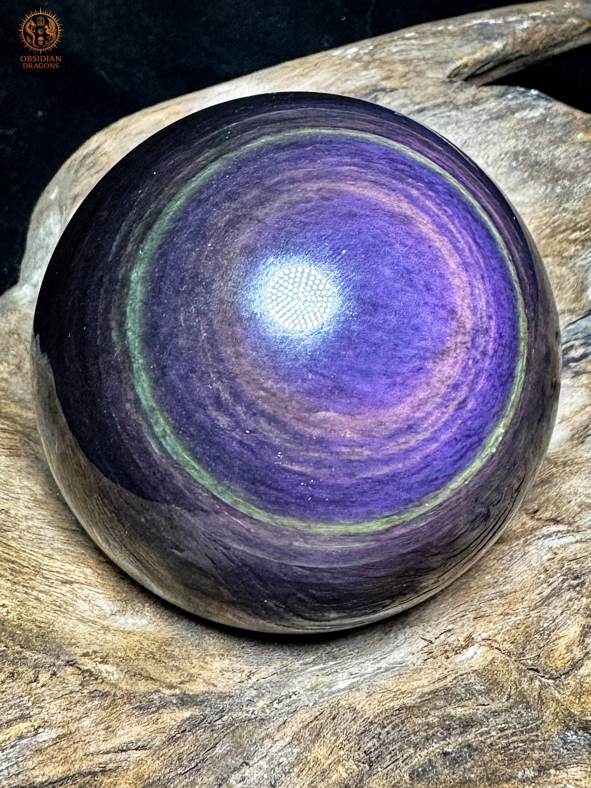 Sphère en obsidienne Manta Huichol aux irisations bleu-violet, couronnes lavande et mince anneau vert, présentée sur bois.