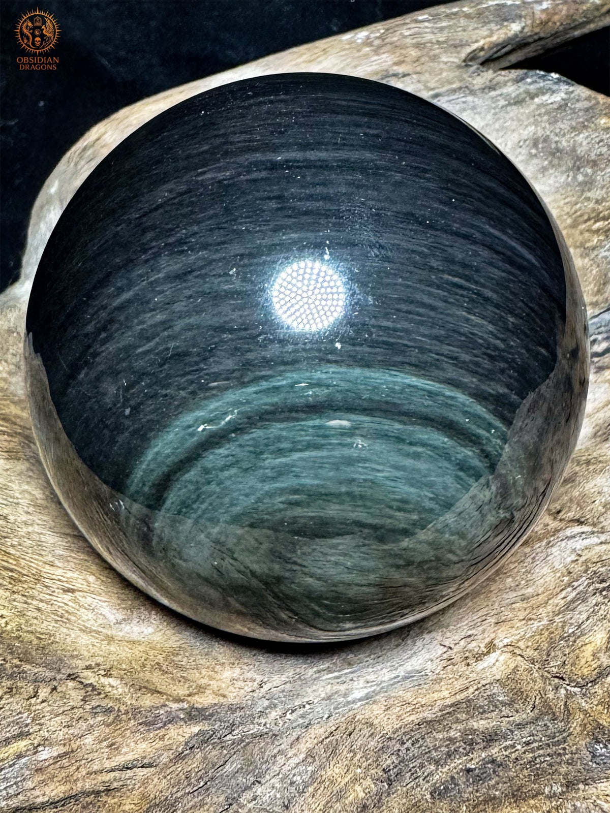 Vue latérale sombre de la sphère en obsidienne Manta Huichol, fines bandes grisées et couronnes vertes discrètes dans la partie basse.