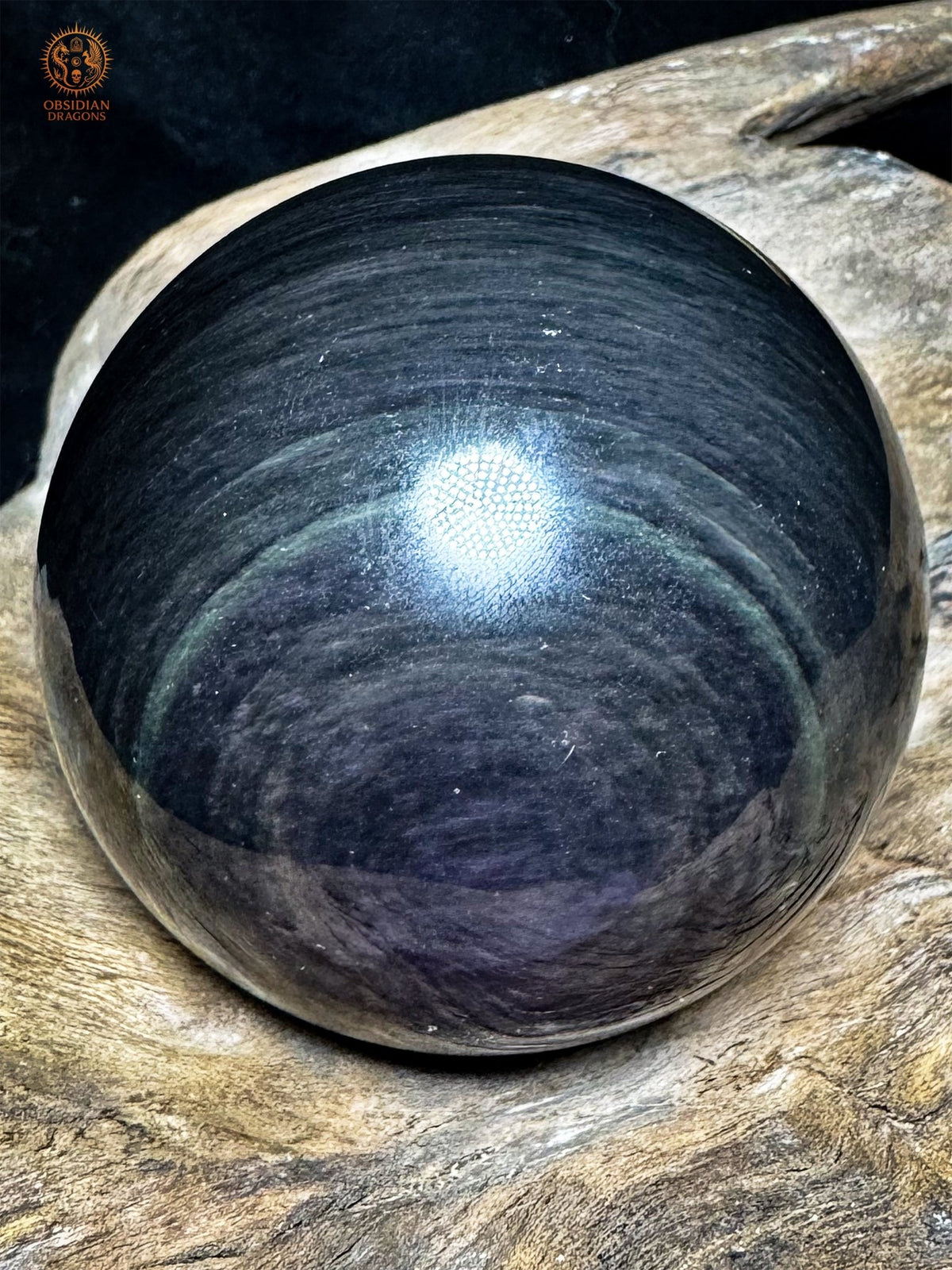 Sphère en obsidienne œil céleste sous un angle sombre, bandes gris-violet et anneau vert discret sur la surface noire réfléchissante.