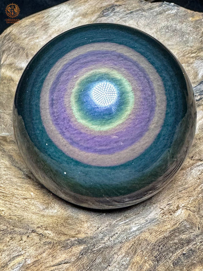 Sphère en obsidienne œil céleste Manta Huichol, aux anneaux concentriques violets et jaune-vert autour d’un centre bleu, posée sur bois - Obsidian Dragons