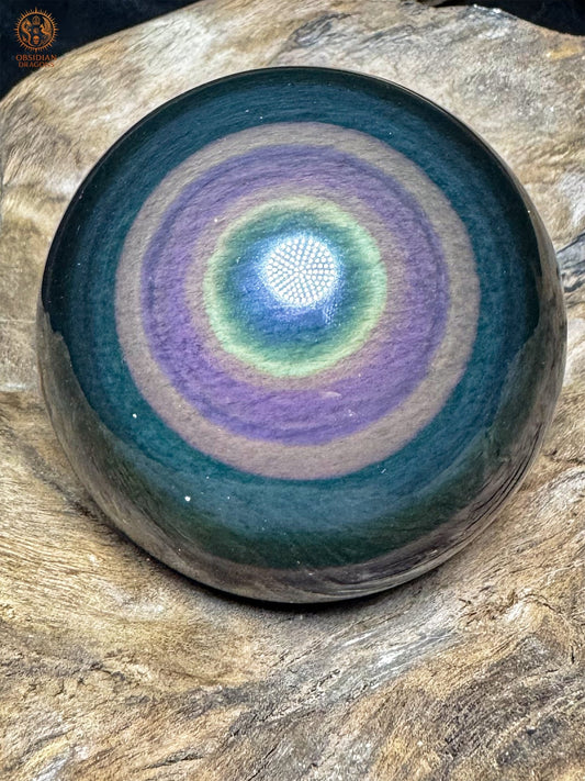 Sphère en obsidienne œil céleste Manta Huichol, aux anneaux concentriques violets et jaune-vert autour d’un centre bleu, posée sur bois - Obsidian Dragons