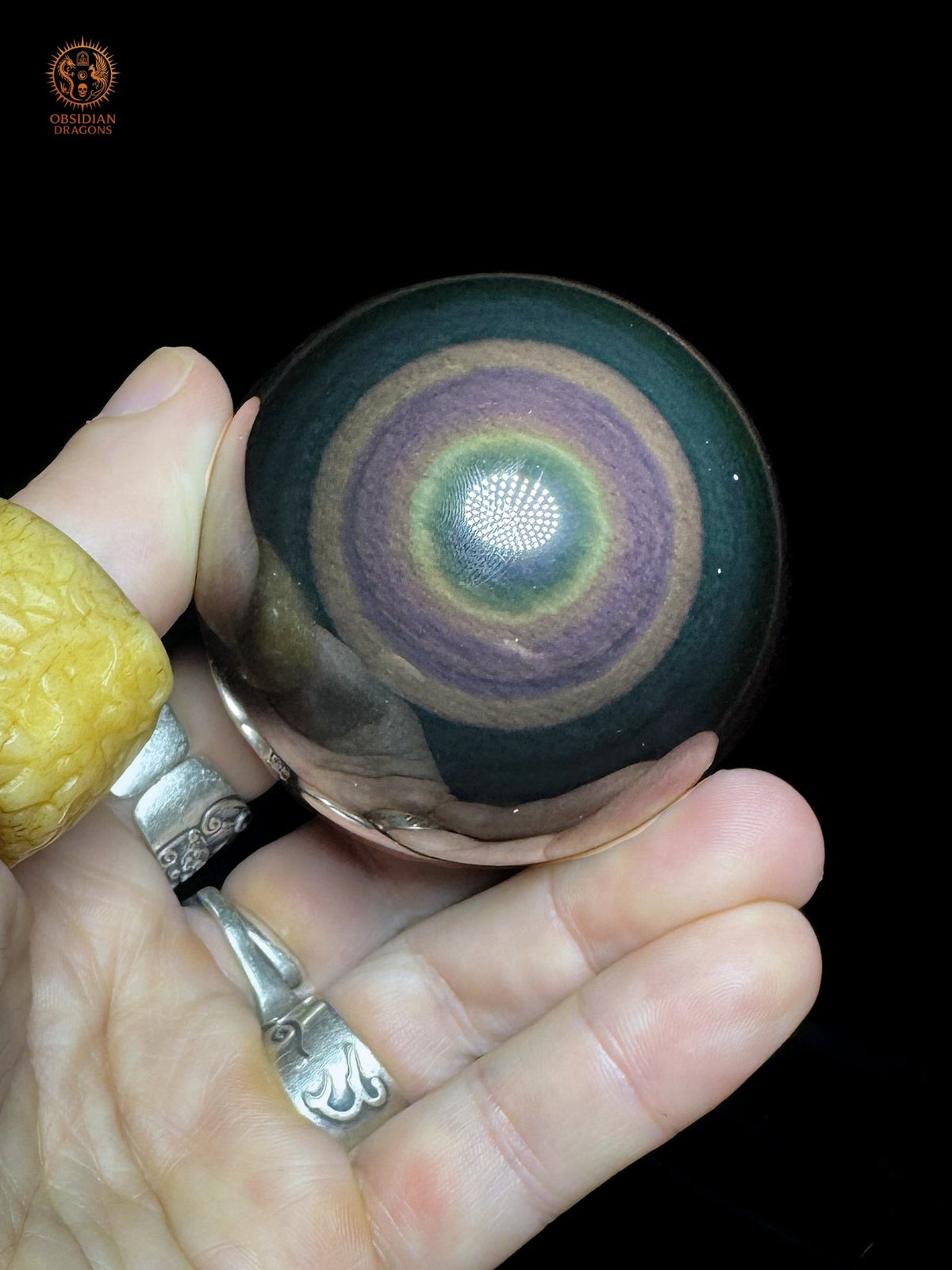 Sphère en obsidienne œil céleste de 6,5 cm tenue en main sur fond noir, montrant des anneaux violets et un halo jaune-vert autour d’un œil bleu - Obsidian Dragons