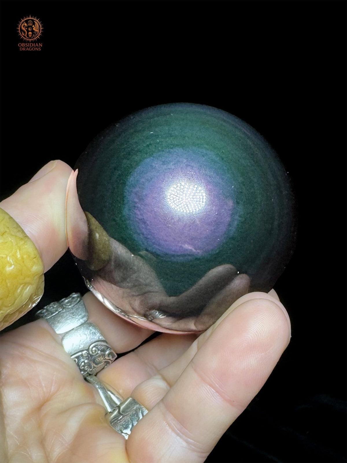 Face panachée de la sphère en obsidienne Manta Huichol, tenue en main sur fond noir, aux reflets violets, bleus et verts - Obsidian Dragons
