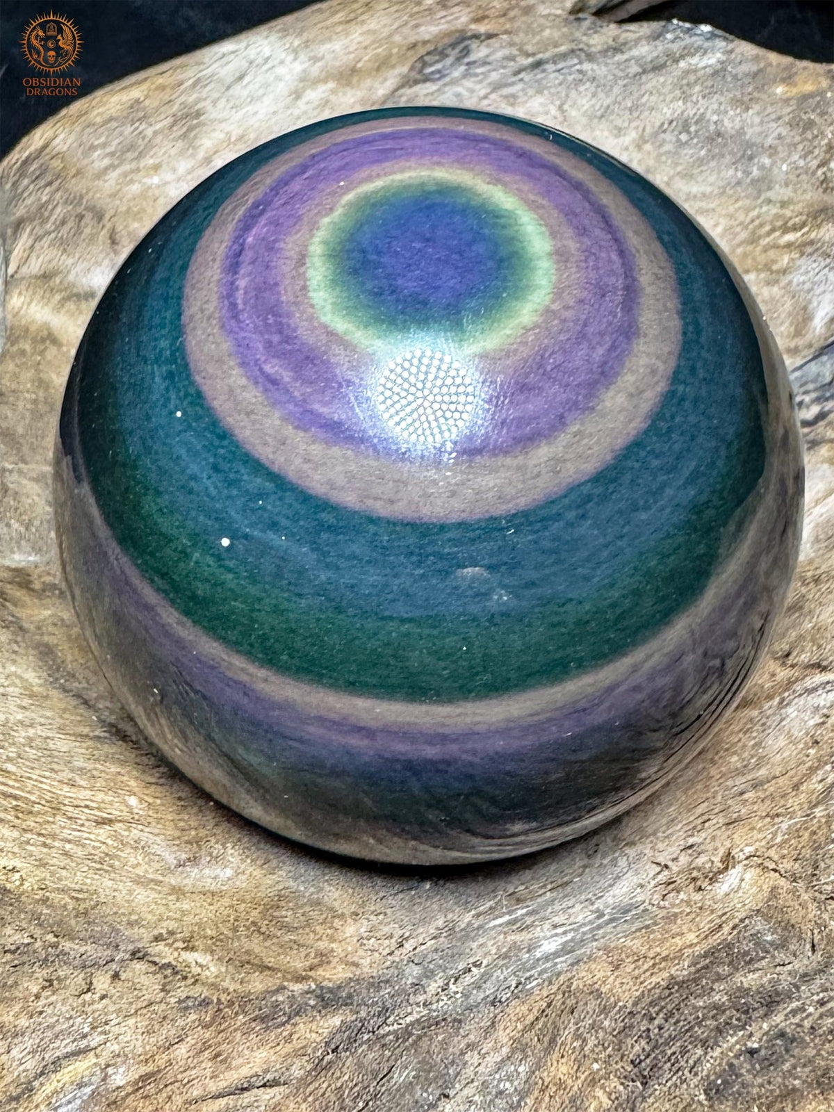 Sphère en obsidienne œil céleste sur bois, avec un œil bleu en partie haute, un halo jaune-vert et de larges couronnes violettes.