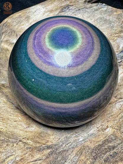 Sphère en obsidienne œil céleste sur bois, avec un œil bleu en partie haute, un halo jaune-vert et de larges couronnes violettes.