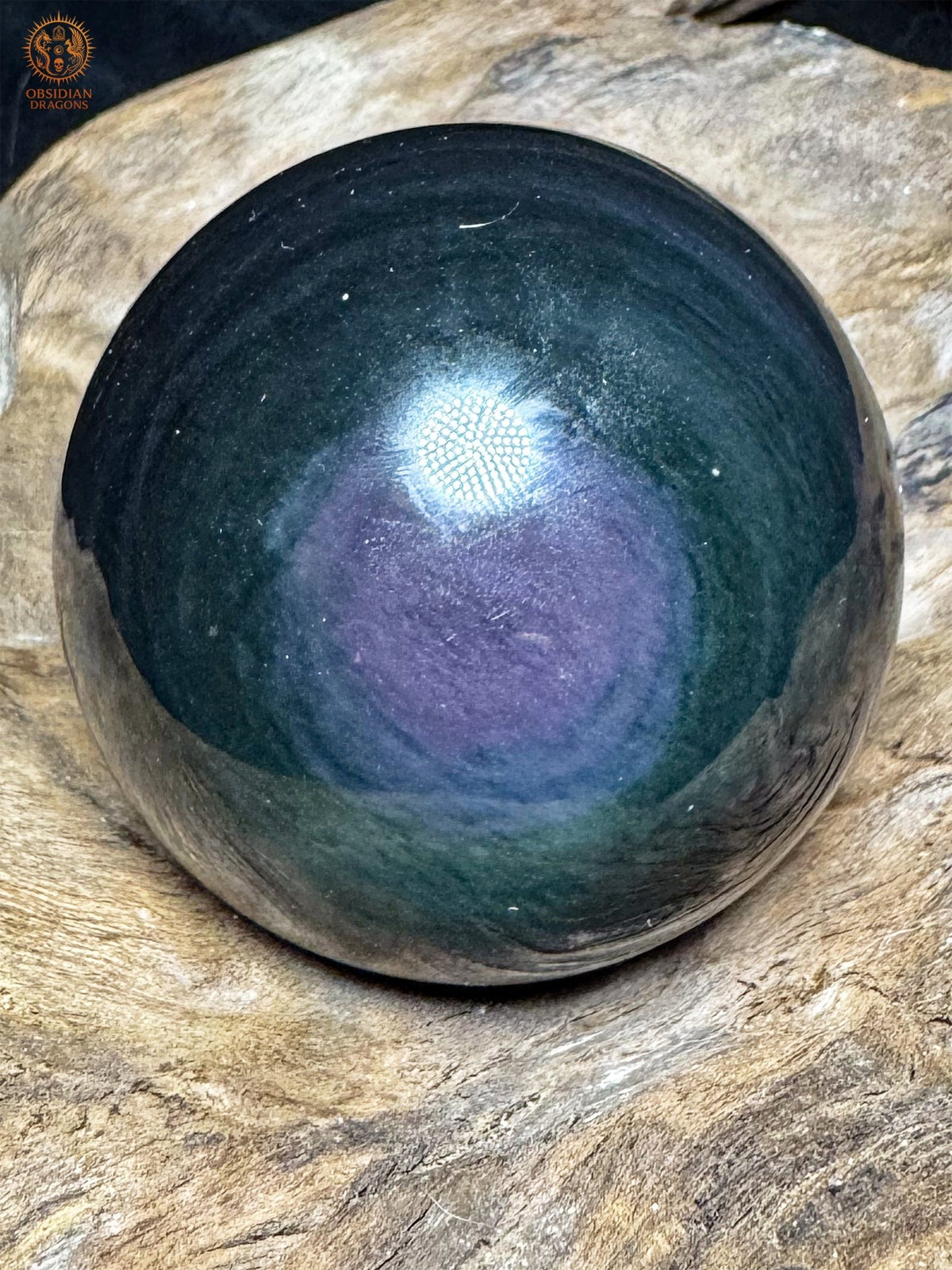 Sphère en obsidienne Manta Huichol posée sur bois, montrant une zone violette décentrée vers le bas, bordée de bleu et de vert sombre.