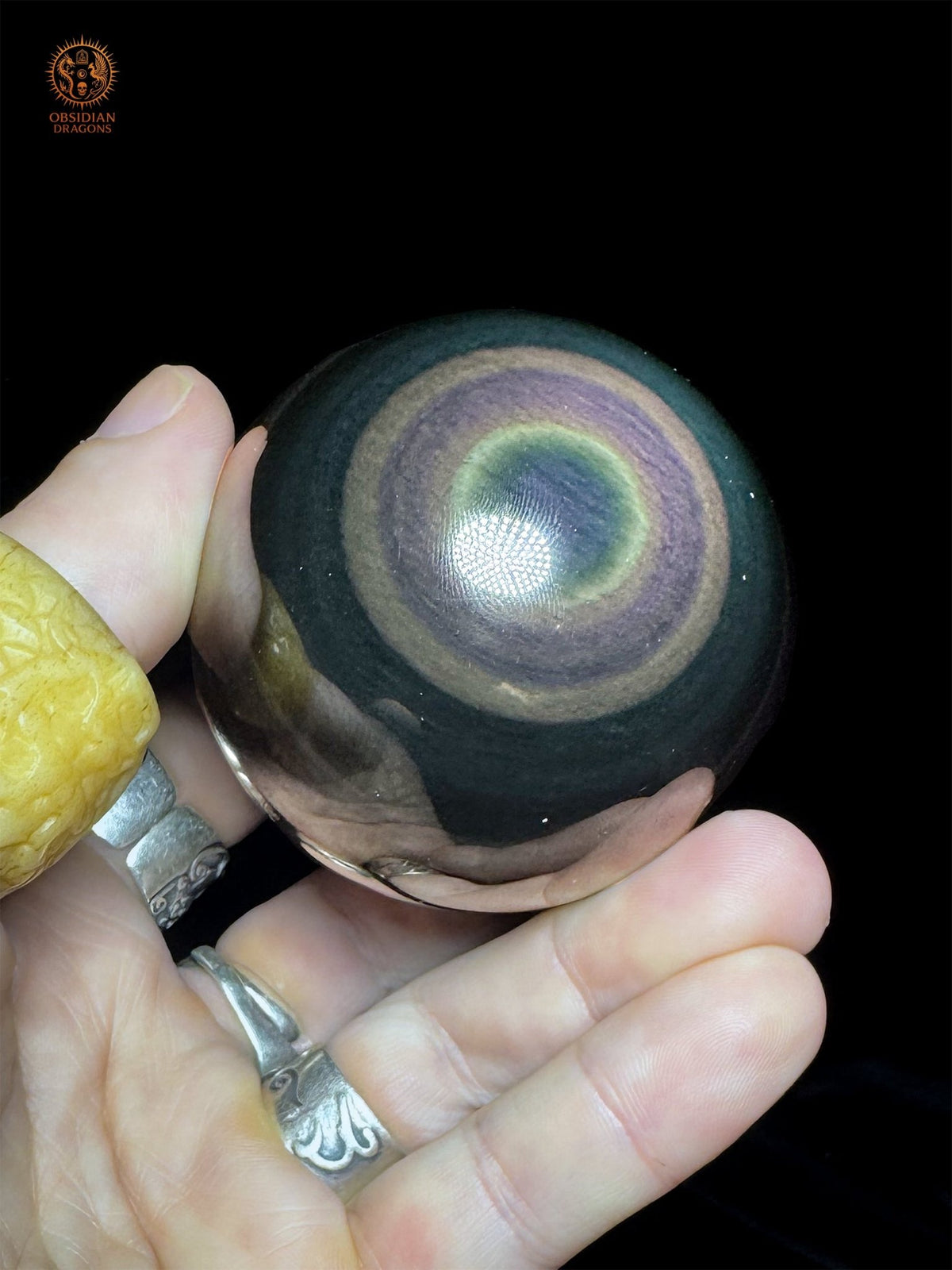 Sphère en obsidienne œil céleste mexicaine tenue dans la paume, avec un œil bleu, un fin halo jaune-vert et des anneaux violets bien délimités.