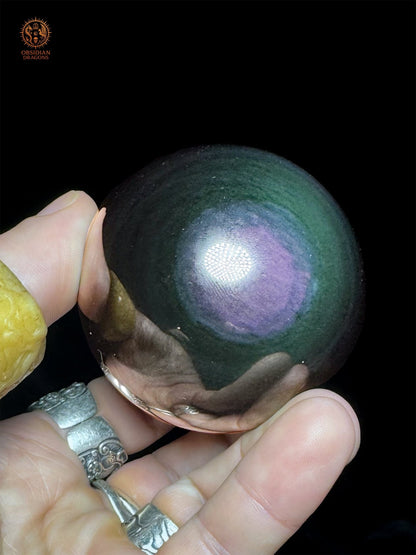 Sphère en obsidienne Manta Huichol de 6,5 cm présentée en main, aux plages violettes et bleues entourées de vert profond, sur fond noir.