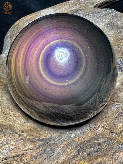 Sphère en obsidienne œil céleste aux reflets mauves, avec un centre violet entouré d’un fin cercle jaune pâle, sur du bois - Obsidian Dragons