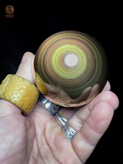 Sphère en obsidienne œil céleste de 7 cm tenue dans la paume, avec un halo jaune-vert et des anneaux concentriques mauves et bronze- Obsidian Dragons