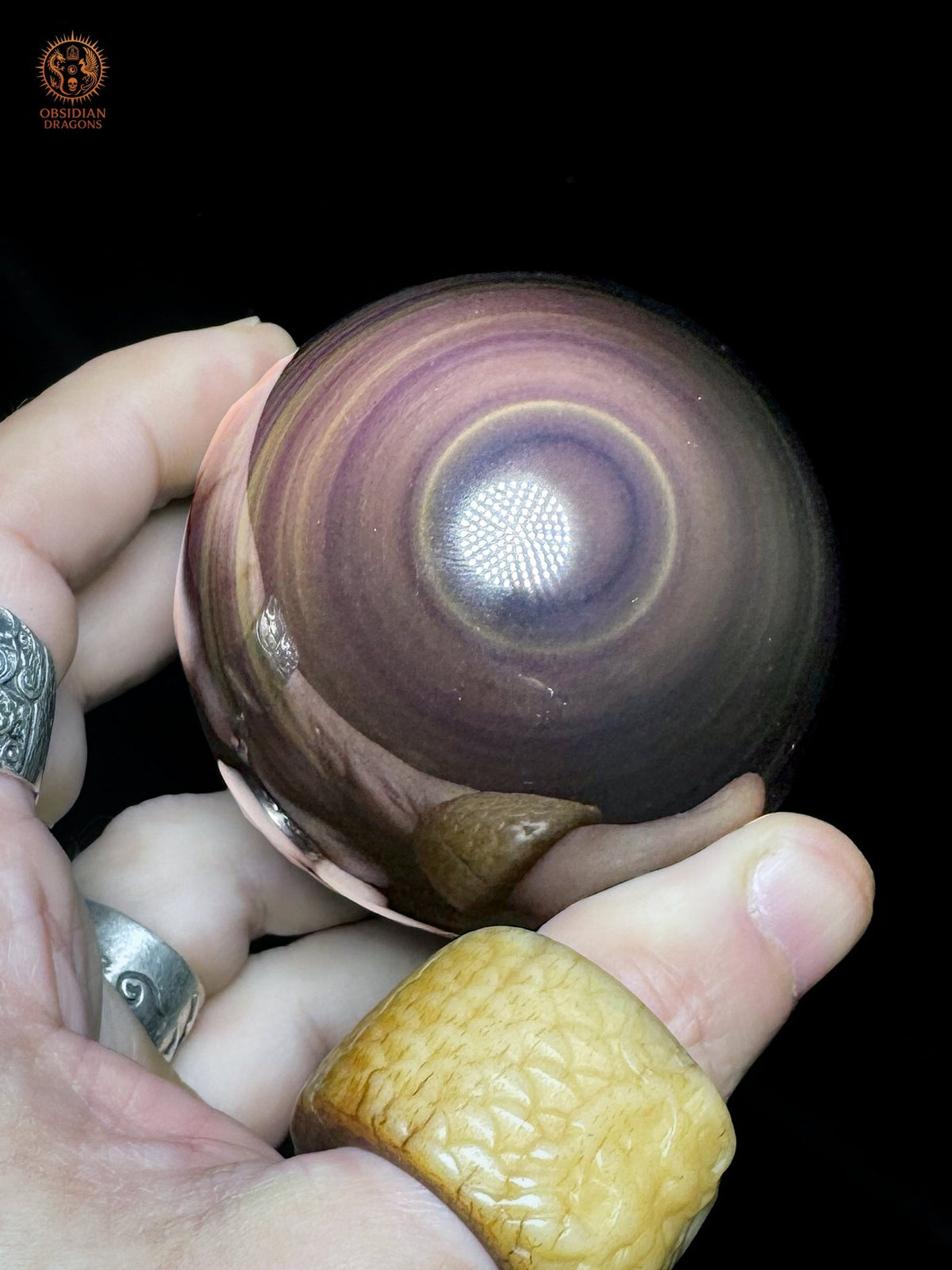 Sphère en obsidienne œil céleste tenue entre les doigts, présentant un œil violet bordé d’un fin cercle jaune et des reflets mauves - Obsidian Dragons