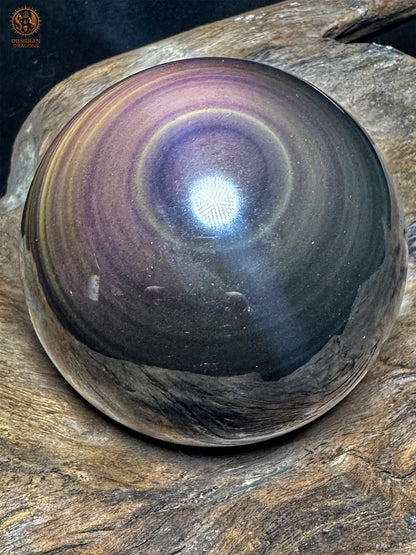 Sphère en obsidienne œil céleste posée sur du bois, avec un centre violet cerclé de jaune pâle, des bandes mauves et une partie inférieure sombre
