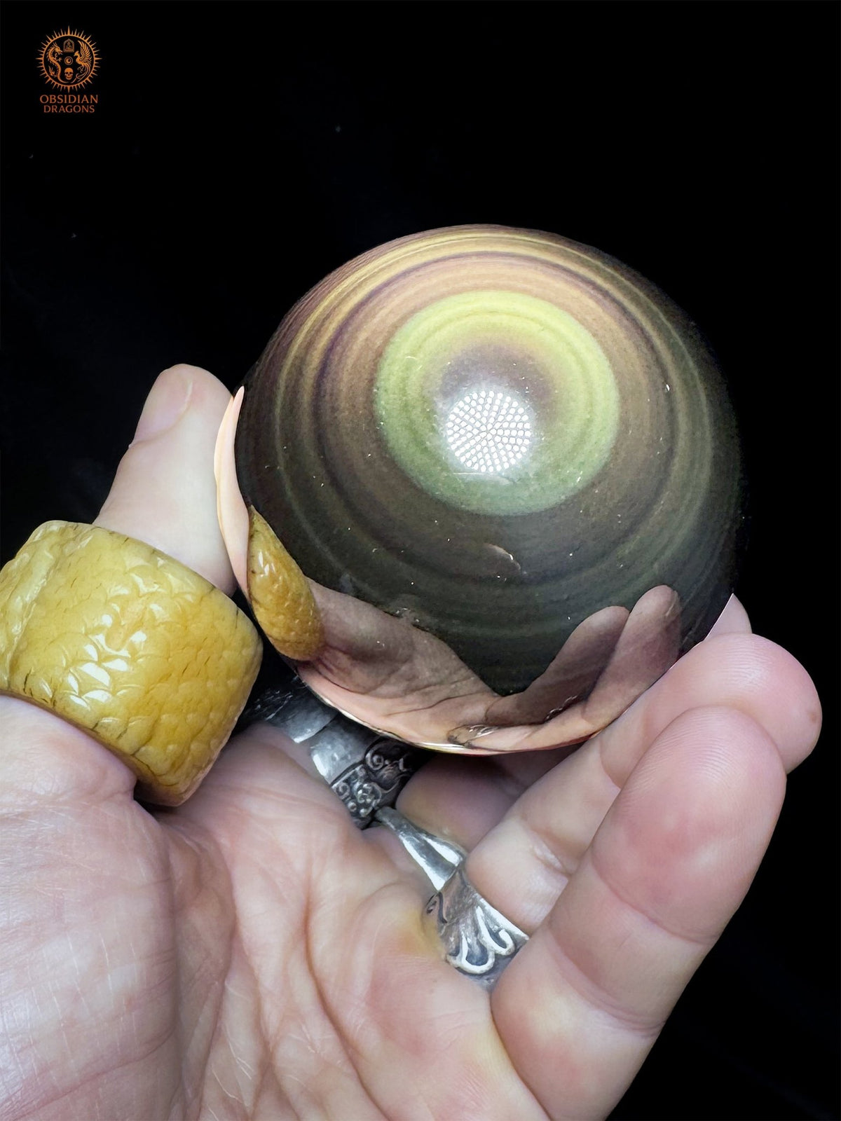 Sphère en obsidienne œil céleste inclinée dans la main, révélant un halo jaune-vert lumineux et des bandes concentriques aux reflets dorés