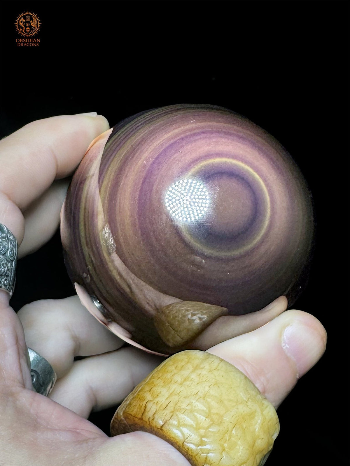 Sphère en obsidienne œil céleste tenue en main, avec un œil violet incliné, un fin cercle jaune et de larges anneaux mauves sur sa surface polie