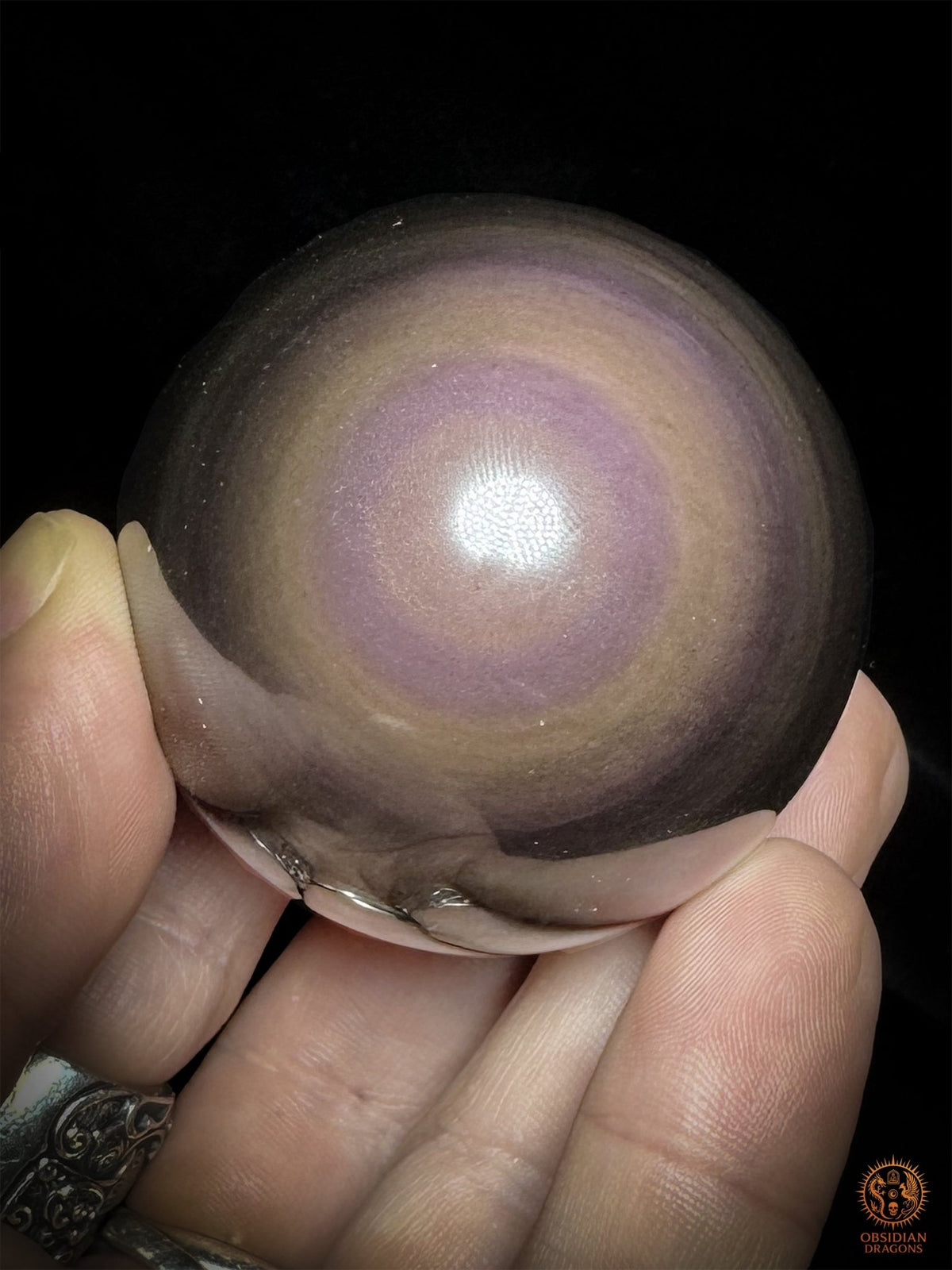 Sphère en obsidienne tenue en main sur fond noir, œil mauve à lavande entouré de couronnes aux nuances rosées et cuivrées - Obsidian Dragons