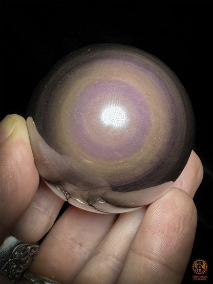 Sphère en obsidienne tenue en main sur fond noir, œil mauve à lavande entouré de couronnes aux nuances rosées et cuivrées - Obsidian Dragons