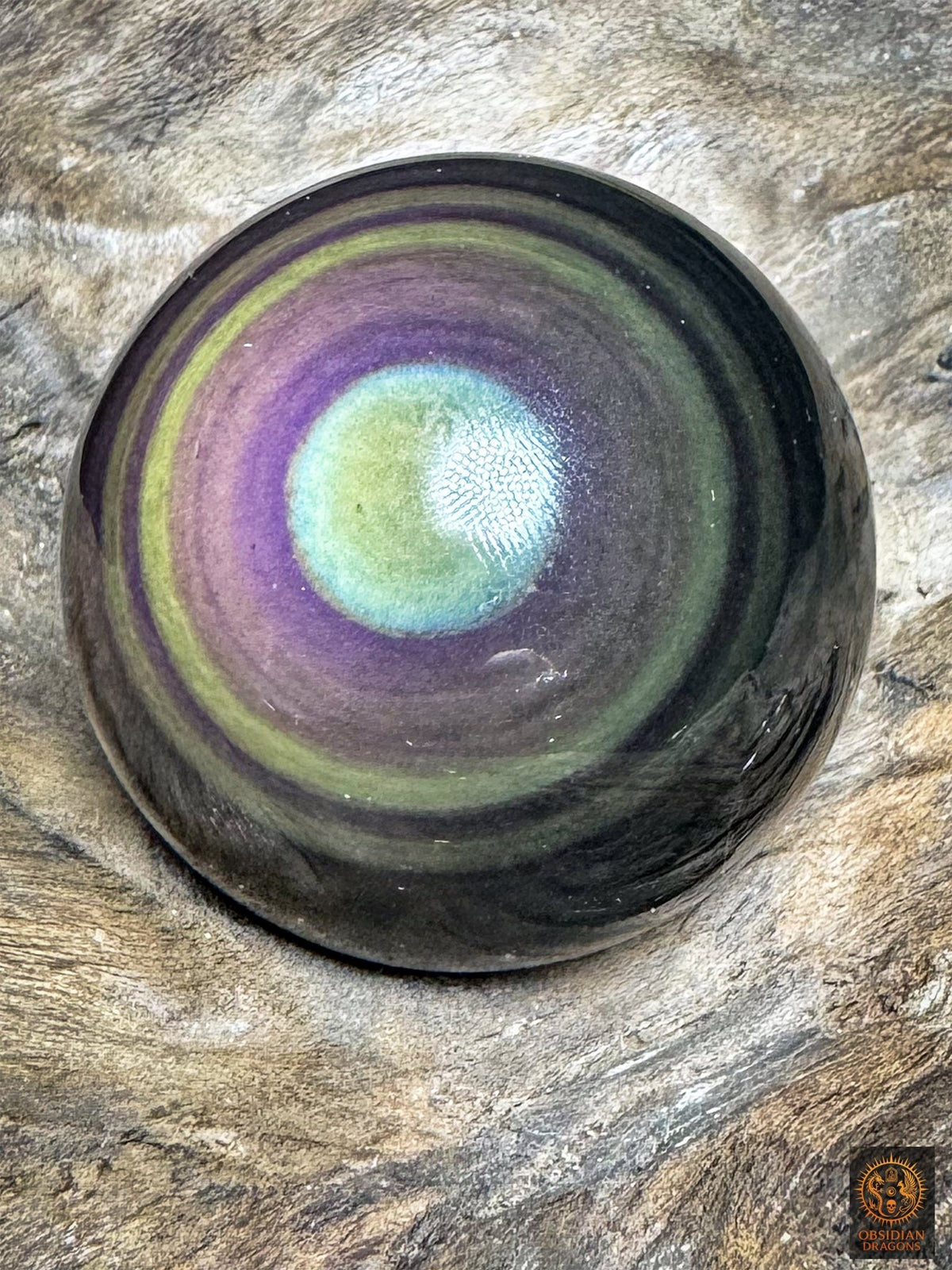Sphère en obsidienne œil céleste vue légèrement de biais, centre vert clair bordé de bleu cyan et anneaux violets et mauves.