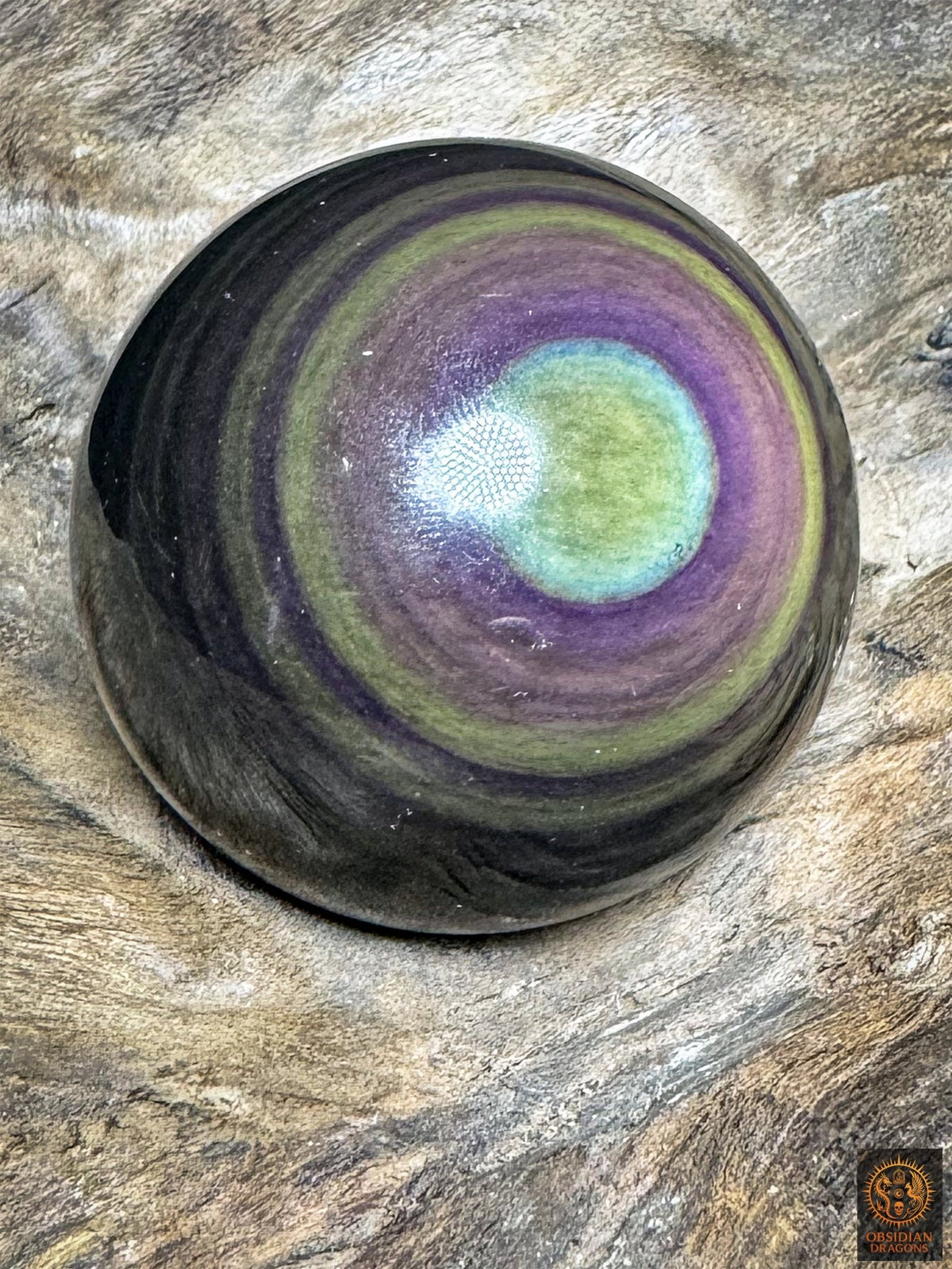 Vue oblique de la sphère en obsidienne, œil jaune-vert bordé de turquoise et entouré de couronnes violettes, mauves et vertes.