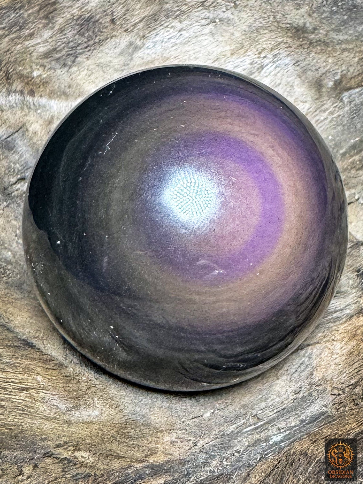 Sphère en obsidienne aux reflets violets et mauves, œil décentré vers la droite et bandes concentriques sur fond noir.